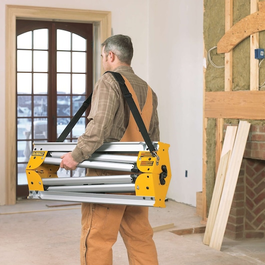 A person wearing work clothes carries a folded DEWALT miter saw stand with a shoulder strap inside a room under construction.