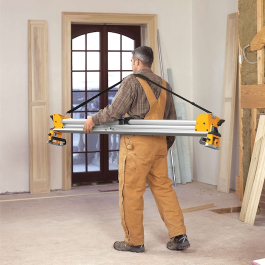 Person wearing work overalls carrying a DEWALT miter saw stand with a shoulder strap inside a room under renovation.