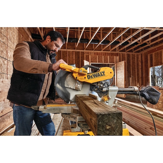 A person operates a DEWALT DWS780 miter saw to cut a wooden beam inside a construction site. The saw is yellow and features the DEWALT logo prominently. The background shows exposed wooden framing and unfinished walls.