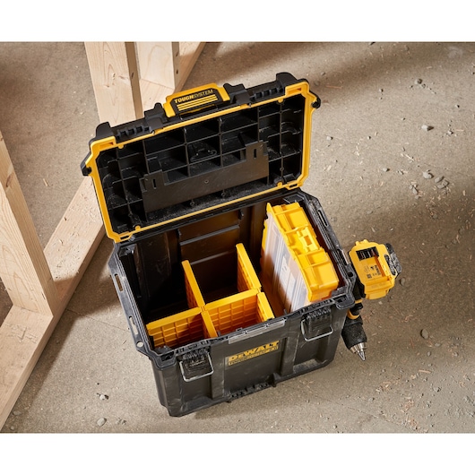 Open DEWALT ToughSystem storage box with yellow compartments and a drill attached on the side, placed on a concrete floor next to wooden framing.