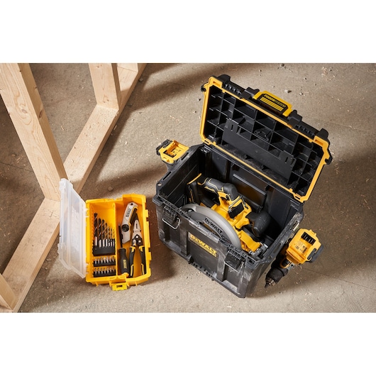 A DEWALT tool storage box with the lid open, containing a circular saw. Next to it is a yellow and transparent smaller organizer with drill bits and pliers, both placed on a workshop floor.