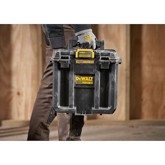 A person wearing work gloves is carrying a DEWALT TOUGHSYSTEM 2.0 black storage box with yellow labels, designed for tool organization and transport, in a construction environment with wooden walls.