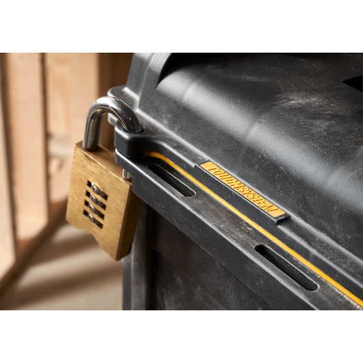 Close-up image of a DEWALT black storage box with a gold combination padlock securing the lid. The box features the DEWALT branding and yellow detailing, and appears to be used in a workshop setting.