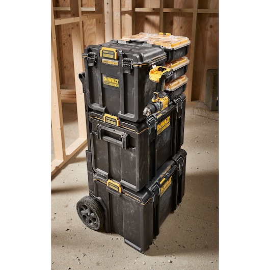 Three stacked DEWALT black and yellow toolboxes with wheels, placed on a construction site floor, with wooden framing in the background.