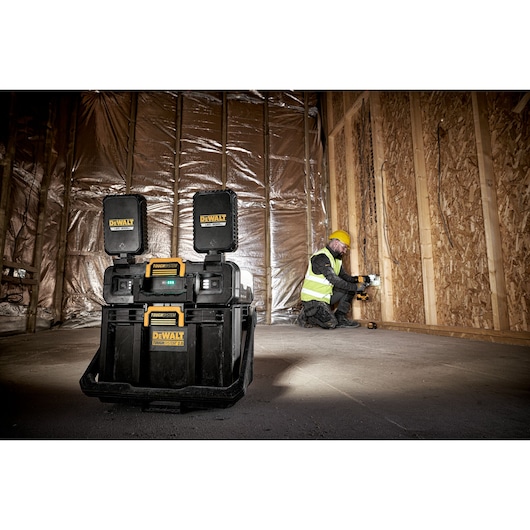 A DEWALT ToughSystem jobsite radio is placed on the floor of a construction site with unfinished wooden walls and insulation. A worker wearing a yellow hard hat and a high-visibility vest is working in the background.