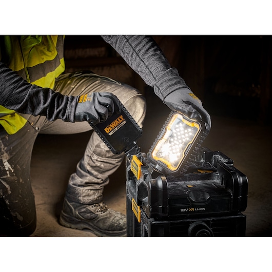 A person wearing work gloves and a yellow safety vest is adjusting a DEWALT portable work light, which is illuminated and placed on top of a black DEWALT tool box. The scene suggests a construction or workshop environment.