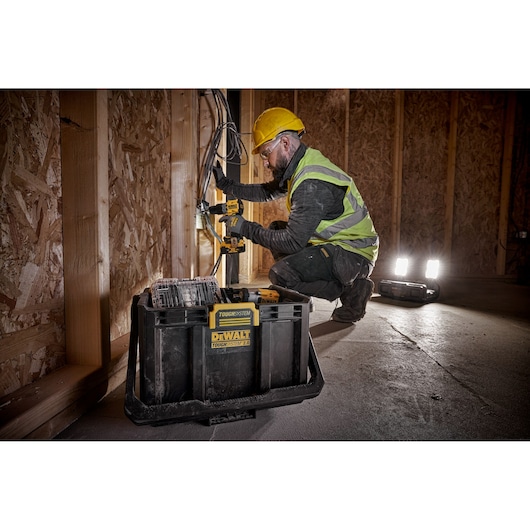 A person wearing a yellow hard hat and a high-visibility vest is using a DEWALT power tool to work on wiring inside a wooden construction site. A DEWALT TOUGHSYSTEM 2.0 toolbox is prominently placed in the foreground, and portable work lights are illuminating the area.