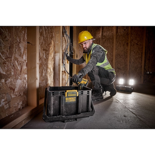 A person wearing a yellow hard hat and safety vest is working with electrical wires inside a wooden framed building. In front of them is a DEWALT ToughSystem storage box, and two portable work lights are placed on the floor.