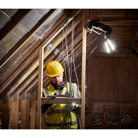 A construction worker wearing a yellow hard hat and high-visibility vest is working in a partially finished attic space. The worker is handling tools and electrical wires, and a portable DEWALT work light is mounted above, illuminating the area.