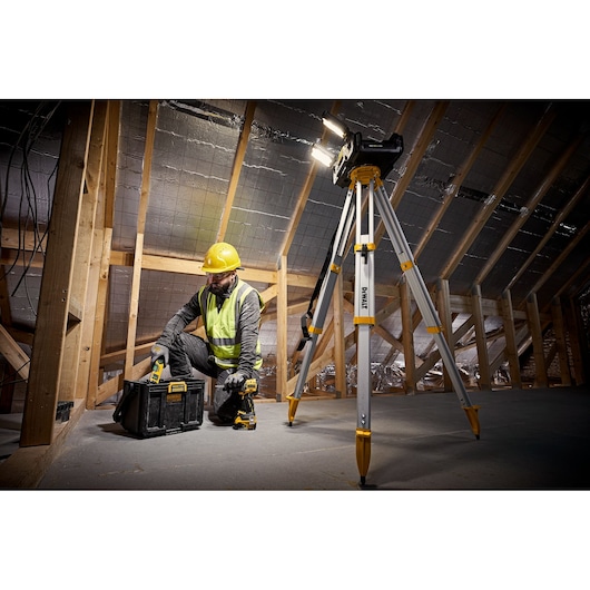 A DEWALT tripod with mounted work light is set up in an unfinished attic. Next to it, a construction worker in safety gear uses a DEWALT toolbox and power tool.