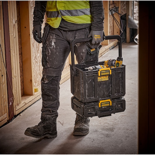 A person wearing work gloves and a high-visibility vest is carrying a DEWALT ToughSystem toolbox filled with tools on a construction site.