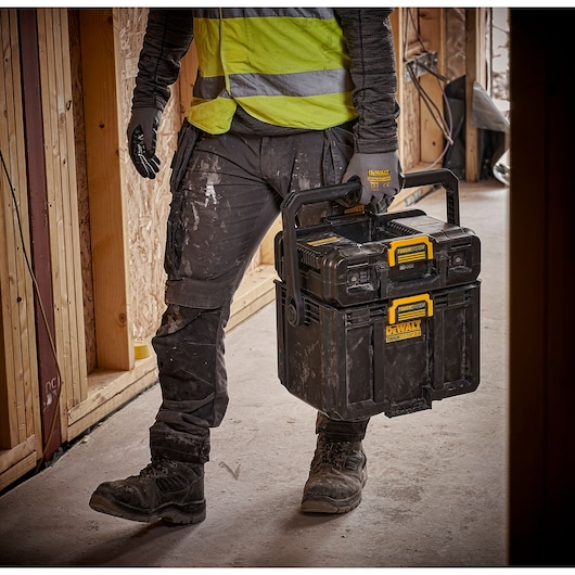 A construction worker wearing a hi-vis vest and gloves carries a DEWALT ToughSystem 2.0 tool box with two stacked storage units at a construction site.
