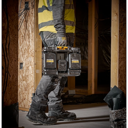 A person wearing a high-visibility vest and work gloves carries a DEWALT DWST08061-1 black and yellow toolbox on a construction site.