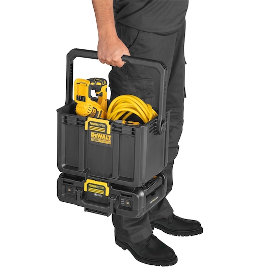 A person holding a DEWALT ToughSystem 2.0 storage box filled with tools and a yellow extension cord. The black and yellow storage box is being carried by its handle, and it sits on a stacked toolbox system. The person is wearing black work pants and shoes.