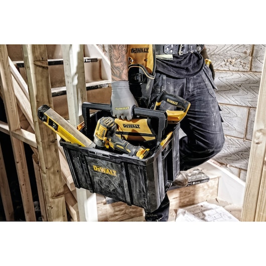 Person holding a DEWALT tool box with tools at a construction site. The box contains a drill and a level.