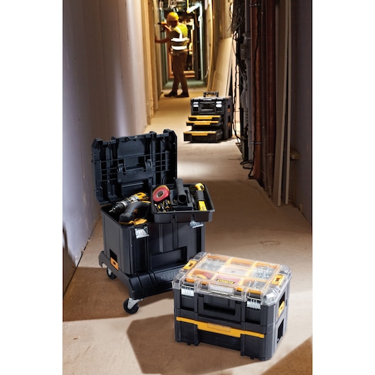 DEWALT DWST1-71229 tool storage boxes with tools, shown in a construction hallway with a worker in the background.