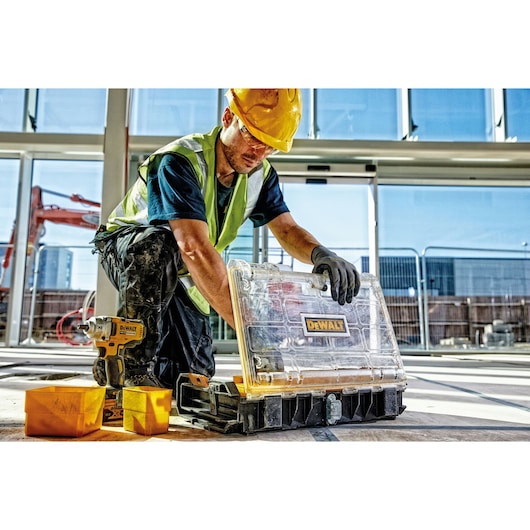 Worker on site with DEWALT tool case, drill, and containers near a window. Safety gear is worn.