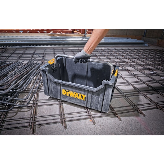 Person lifts a DEWALT plastic tool box with a handle, placed on a metal grid in a construction site.
