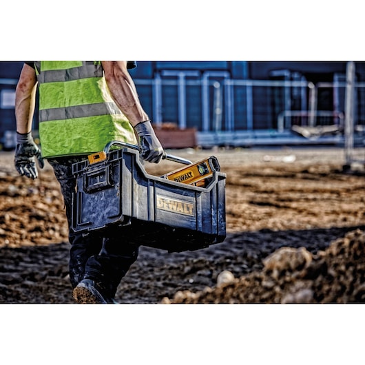 Person carries a DEWALT toolbox with tools on a construction site, wearing gloves and a reflective vest.