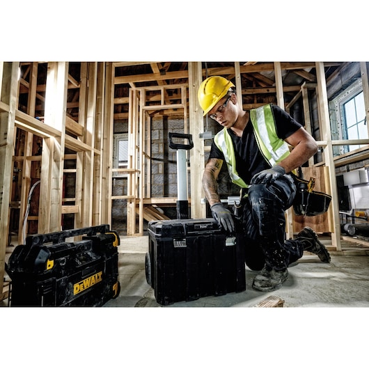 Worker kneeling in a construction site with DEWALT toolboxes. Wooden framing and building materials are visible.