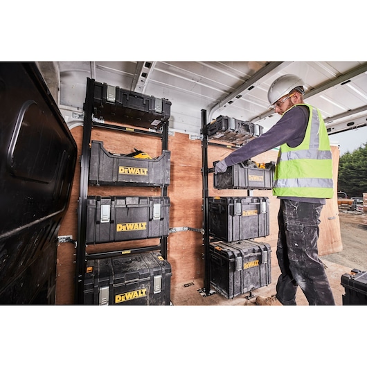 Worker stores DEWALT toolboxes on rack inside van; wearing safety gear. Toolboxes organized on shelves.