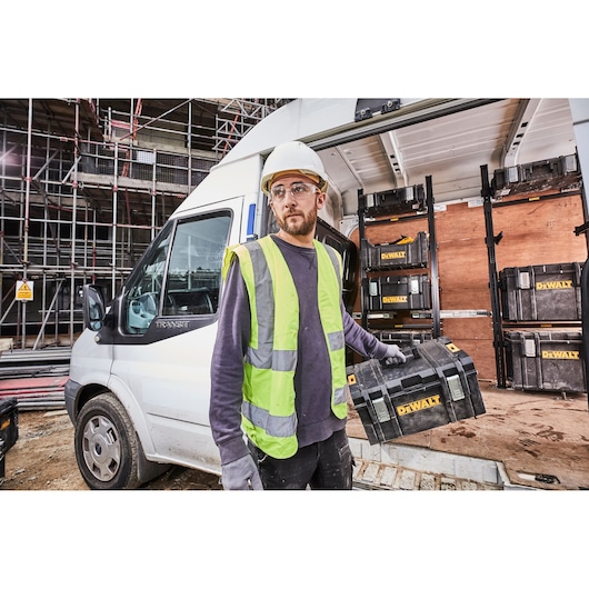 Worker in hi-vis holding a DEWALT toolbox in front of van with more DEWALT toolboxes inside, at a construction site.