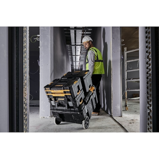 Worker pulls stacked DEWALT toolboxes on wheels at construction site in a narrow hallway.
