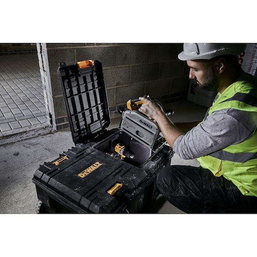 Worker opening a DEWALT toolbox with power tools inside, on a construction site with a concrete wall.