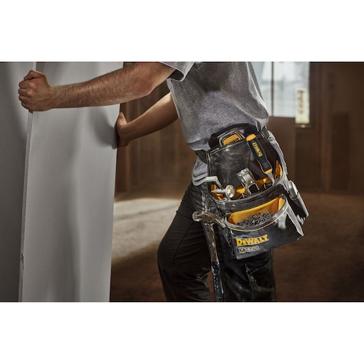 A person wearing a DEWALT tool belt holds a large panel. The tool belt contains various hand tools, including a hammer, screwdrivers, and fasteners. The scene is set indoors, suggesting a construction or installation environment.