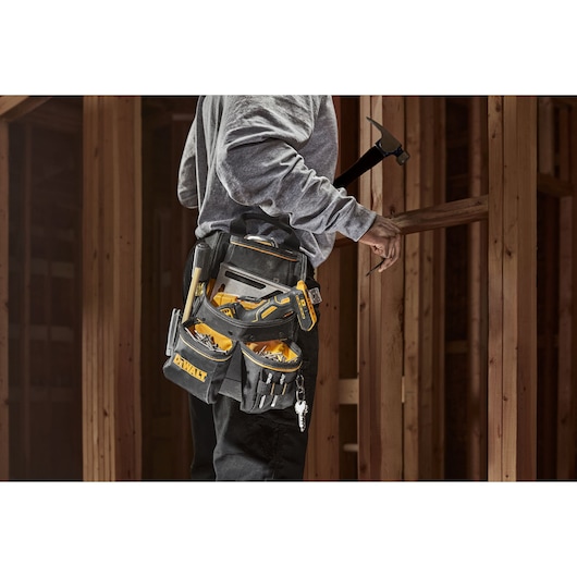 A person wearing a DEWALT tool belt loaded with various hand tools, working in a wooden structure. The tool belt has multiple pockets and compartments for organized tool storage.