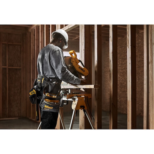 Person wearing a hard hat and tool belt operating a DEWALT miter saw in a wooden frame construction site.