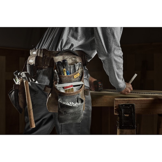 A close-up of a person wearing a DEWALT tool belt loaded with various tools such as a hammer, pencils, and screwdrivers. The person is working at a wooden table and marking a piece of wood.