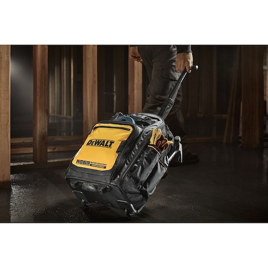 A DEWALT branded black and yellow rolling tool bag with an extendable handle, being pulled by a person in a worksite environment. The bag is equipped with exterior pockets holding work gloves and other tool accessories.