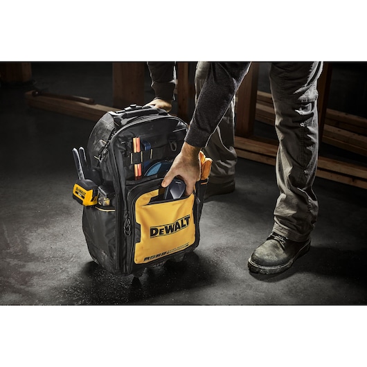 A person is holding a DEWALT branded backpack tool bag, which is filled with various tools and equipment. The bag is yellow and black, and is positioned on a workshop floor.