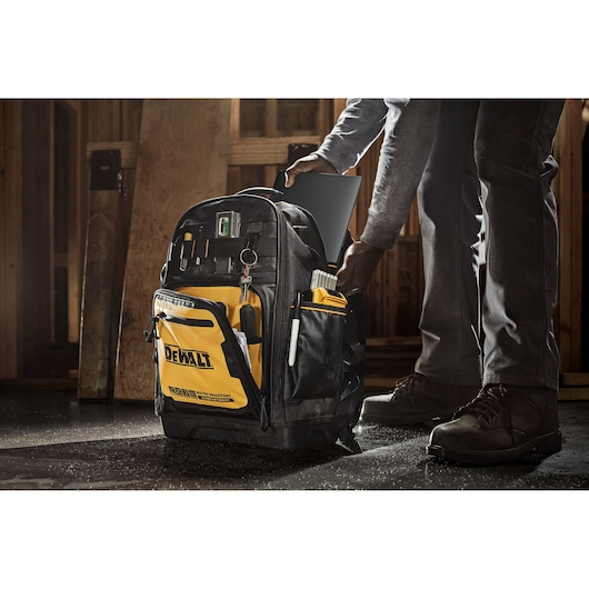 A person places a laptop into a DEWALT tool backpack, which features multiple pockets, organization compartments, and a prominent yellow and black design, standing on a workshop floor.
