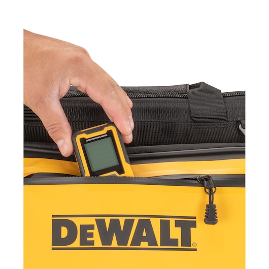 A hand placing a laser distance meter into the zippered pocket of a yellow DEWALT bag.