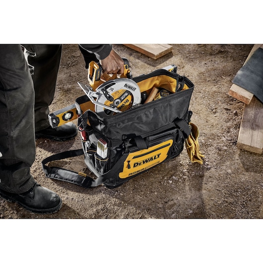 A DEWALT tool bag placed on a construction site floor, filled with various DEWALT tools and accessories including a circular saw, a level, and other hand tools. A person is reaching into the bag to take out the circular saw.