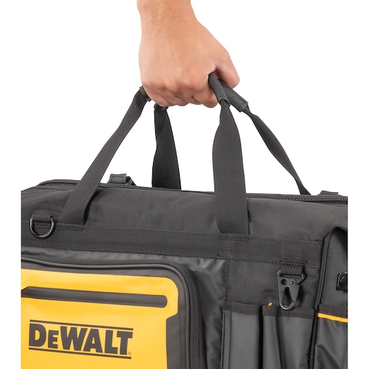 A close-up of a hand holding the handles of a black and yellow DEWALT tool bag with zippered compartments and pockets.