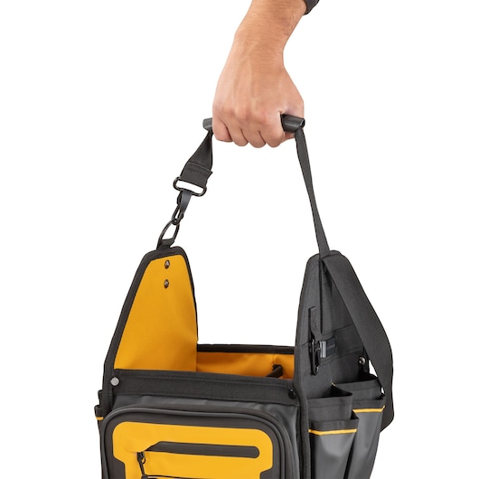 Close-up of a hand holding a DEWALT tool bag with black and yellow colors, showing its handle, open compartments, and external pockets.