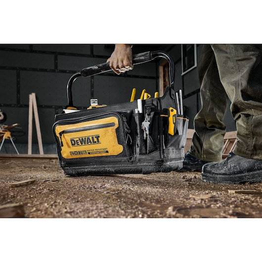A DEWALT-branded black and yellow tool bag, filled with hand tools, is being carried by a person on a construction site with a dirt floor.