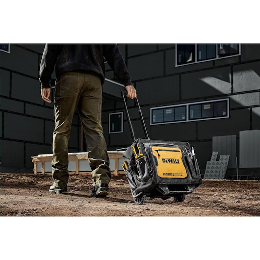 A person is pulling a DEWALT rolling tool bag with a telescopic handle across a dirt construction site. The bag is black and yellow, labeled with the DEWALT logo, and has several compartments for storage.