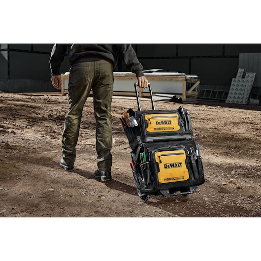 A person is pulling a DEWALT rolling tool bag on a construction site. The bag is black and yellow with multiple pockets and compartments, and is filled with various tools.