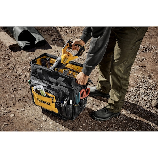 A person placing a yellow DEWALT power tool into a black and yellow DEWALT tool bag filled with various tools and accessories, on a dirt ground at an outdoor work site.