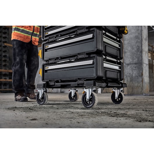 Stacked DEWALT toolboxes on wheels in a construction area, shown near a worker's legs.