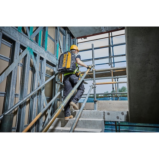 Worker climbs stairs at a construction site carrying a yellow DEWALT backpack.