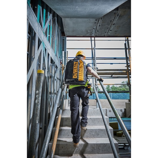 Worker with DEWALT backpack climbing stairs at a construction site, wearing a yellow helmet and safety gear.