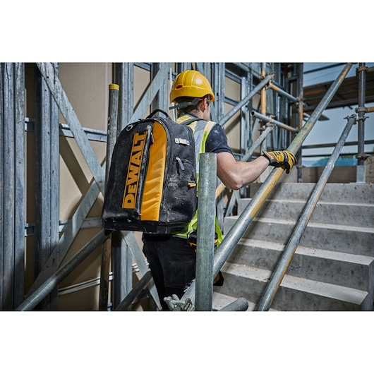 Worker with DEWALT backpack ascending stairs on a construction site, wearing safety gear and helmet.