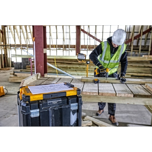 A DEWALT TSTAK toolbox with yellow and black design is placed in the foreground of a construction site. On top of the toolbox is a sheet of paper with diagrams. In the background, a construction worker wearing a hard hat and safety vest is working with a power tool on wooden planks.