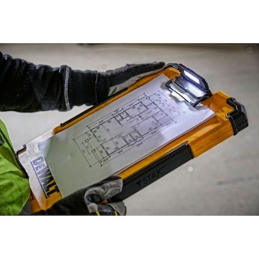 A person wearing gloves holds a DEWALT TSTAK clipboard with an integrated LED light, displaying a technical floor plan drawing attached to the clip.