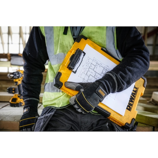 A person wearing work gloves and a high-visibility vest is holding a yellow DEWALT clipboard case with architectural drawings attached, at a construction site.